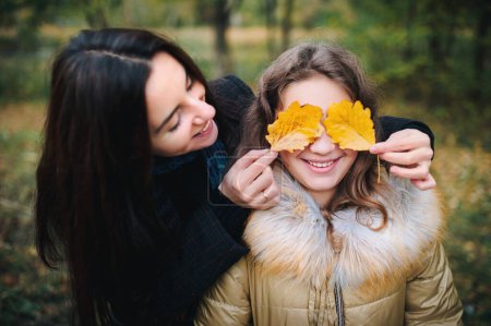 Téléchargez les photos : Une jeune mère couvrit les yeux de sa fille de feuilles jaunes d'automne. Plaisir dans la forêt d'automne. - en image libre de droit