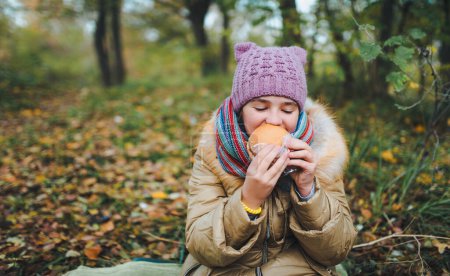 Photo for Portrait of a hungry teen girl, sitting on the ground and eating burger, enjoying picnic in the autumn forest. - Royalty Free Image