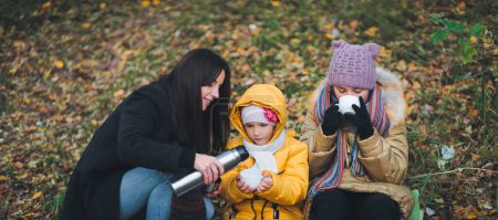 Foto de En una caminata de otoño, mamá vierte té caliente de su termo a sus hijas. El concepto de un paseo en familia. - Imagen libre de derechos