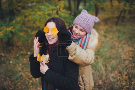 Foto de La hermana menor cerró los ojos de su hermana mayor con hojas amarillas de otoño. Las novias se divierten en el bosque de otoño. - Imagen libre de derechos