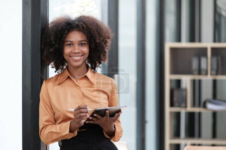 Téléchargez les photos : Heureuse femme d'affaires africaine bavardant sur des gadgets numériques en ligne, tenant un téléphone mobile, utilisant un ordinateur portable, regardant la caméra, souriant. Un employé indépendant blessé à la tête sur son lieu de travail. Haute qualité - en image libre de droit