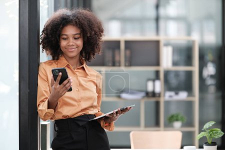 Photo for Happy African business woman chatting on digital gadgets online, holding mobile phone, using laptop computer, looking at camera, smiling. Freelance employee at home workplace head shot. High quality - Royalty Free Image