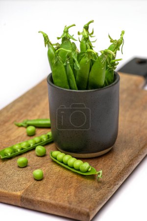 Photo for Green pea pods in a ceramic mug and scattered on a wooden board on white table - Royalty Free Image