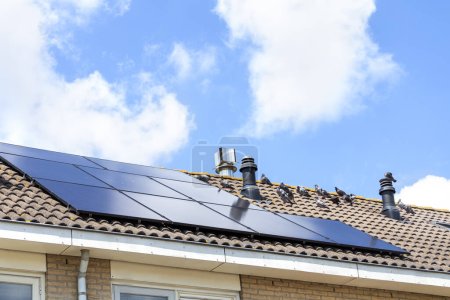 Téléchargez les photos : Batterie solaire sur le toit de la maison sur fond de ciel bleu avec des nuages légers. Utilisation de sources d'énergie alternatives. - en image libre de droit