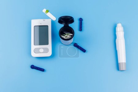 Digitales Glukometer, Lanzettstift, Einmalnadeln und Teststreifen auf pastellblauem Hintergrund. Ansicht von oben. Diabetes-Konzept