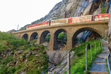 Red train passing by Devil's bridge at St. Gotthard pass on the Swiss Alps in Andermatt, Switzerland