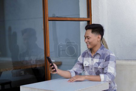 Photo for Young Asian man holding cellphone, sitting in a cafe, viewed from the left side - Royalty Free Image
