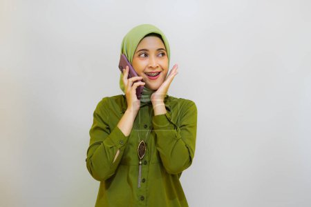 Smiling young Asian Muslim woman dressed in casual shirt talking on mobile phone and looking at copy space isolated over white background