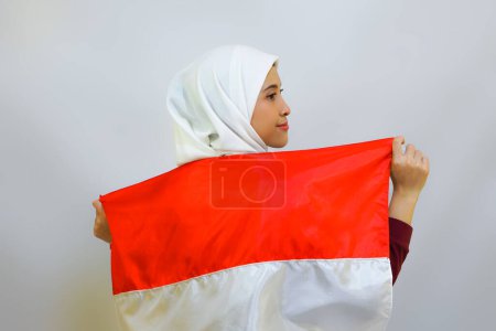 Indonesian muslim woman wearing hijab with proud gesture while holding Indonesia's flag. Indonesia's independence day concept