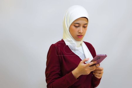 Feliz y alegre joven musulmana asiática vestida con top rojo y hiyab blanco, sosteniendo su teléfono, aislada por el fondo blanco. Concepto del día de la independencia de Indonesia