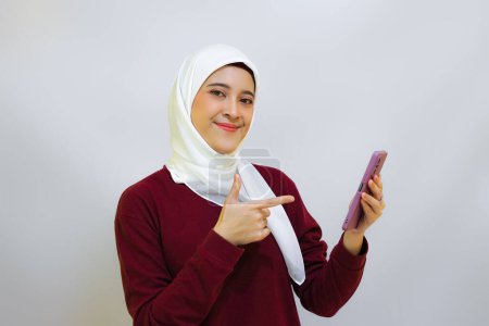 Photo for Happy and cheerful young Asian muslim woman wearing red top and white hijab, holding her phone, isolated by white background. Indonesia's independence day concept - Royalty Free Image