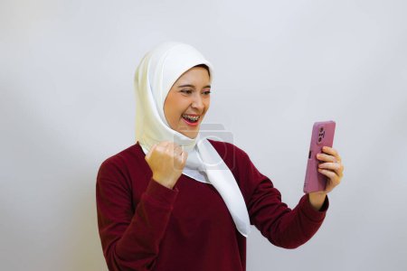 Fröhliche und fröhliche junge muslimische Asiatin mit rotem Oberteil und weißem Hijab, die ihr Handy hält, isoliert von weißem Hintergrund. Indonesiens Konzept zum Unabhängigkeitstag