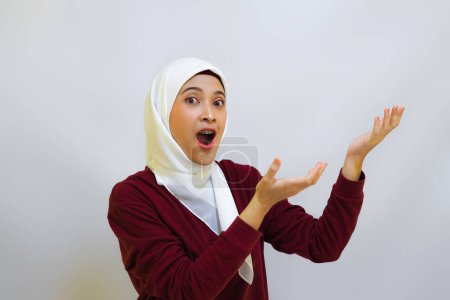 Une musulmane asiatique souriante portant un haut rouge et un hijab blanc, pointant vers sa gauche, isolée par un fond blanc. Concept de fête de l'indépendance de l'Indonésie
