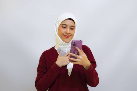 Happy and cheerful young Asian muslim woman wearing red top and white hijab, holding her phone, isolated by white background. Indonesia's independence day concept