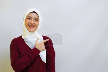 A smiling Asian muslim woman wearing red top and white hijab, pointing to her left, isolated by white background. Indonesia's independence day concept
