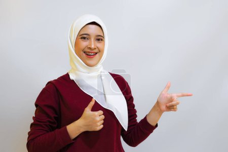 A smiling Asian muslim woman wearing red top and white hijab, pointing to her left, isolated by white background. Indonesia's independence day concept