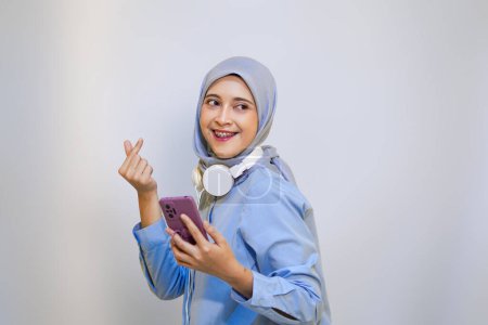 Foto de Mujer alegre musulmán con el dedo signo de amor ama la música en su teléfono móvil. auriculares con cable y el concepto de escuchar música - Imagen libre de derechos