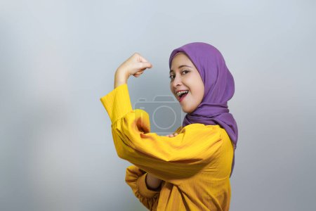 Téléchargez les photos : Côté jeune asiatique musulmane femme porter tenue jaune & hijab montrer biceps muscles sur la main démontrer la force puissance isolé sur fond blanc. - en image libre de droit