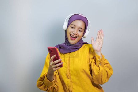 Foto de Chica musulmana feliz en auriculares inalámbricos escuchando música en línea en el teléfono inteligente, disfrutando de una nueva aplicación de fondo blanco - Imagen libre de derechos