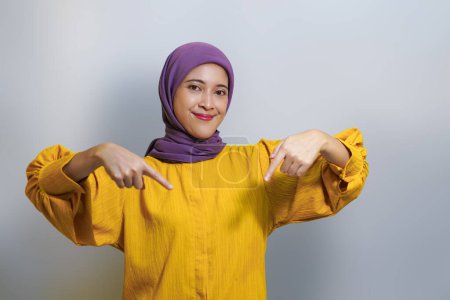 Retrato de alegre hermosa joven musulmana asiática usando hijab y vestido amarillo apuntando con el dedo hacia abajo, mirando a la cámara aislada sobre fondo blanco del estudio