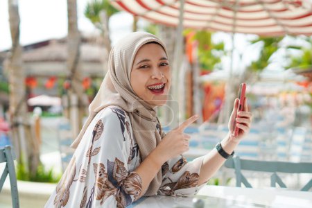 Photo for Woman in Hijab Using Phone. A depiction of a woman wearing a hijab engaged with her mobile device at the beach lounge. - Royalty Free Image