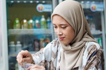 Téléchargez les photos : Hijab indonésien femme versant une boisson emballée dans une tasse avec glaçons au café ou au marché minimal - en image libre de droit