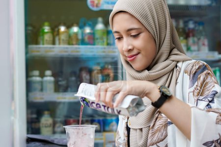 Hijab indonésien femme versant une boisson emballée dans une tasse avec glaçons au café ou au marché minimal