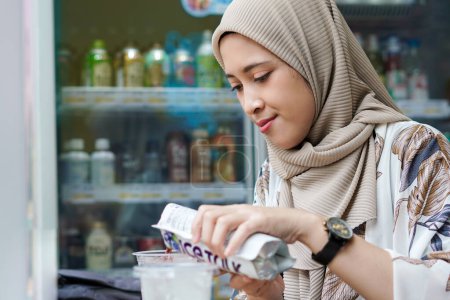Téléchargez les photos : Hijab indonésien femme versant une boisson emballée dans une tasse avec glaçons au café ou au marché minimal - en image libre de droit