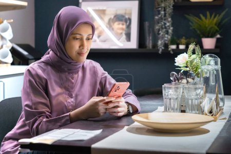 Frau im Hijab sitzt am Esstisch und spielt mit Handy, zeigt zerstreuten oder lächelnden Gesichtsausdruck, als warte sie auf jemanden