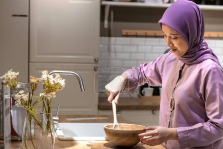 Hübsche muslimische Asiatin im Hijab trägt ein lila Kleid und macht Teig in einer modernen Küche mit Pendellicht und in der Nähe der Spüle