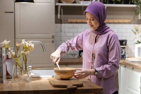 Pretty muslim Asian woman in hijab wearing a purple dress, making dough in a modern kitchen with pendant light and near the sink
