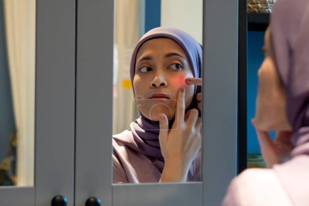 Muslimische Frau sieht sich selbst im Spiegel an und hält ihre Wange wegen Akne