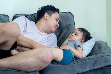 Asian man lying on sofa in living room with little girl on top, both smiling, father and daughter relationship.