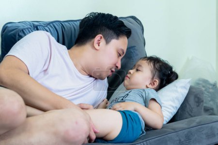 Asiatique homme couché sur canapé dans le salon avec petite fille sur le dessus, souriant, père et fille relation.