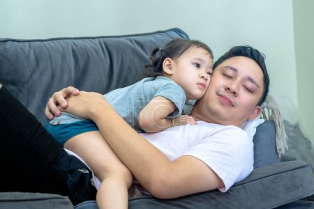 Asiatischer Mann liegt auf Sofa im Wohnzimmer mit kleinem Mädchen an der Spitze, beide lächeln, Vater-Tochter-Beziehung.