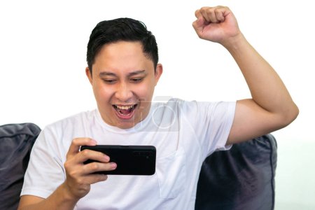Adult Asian man in white t-shirt sitting on sofa, holding cellphone with winning gesture.