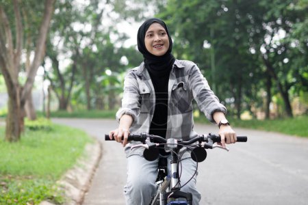 Photo for Young cheerful hijab woman, wearing casual flannel riding bicycle bike on sidewalk in city enjoying being outdoors, look aside. People active urban healthy lifestyle cycling concept. - Royalty Free Image