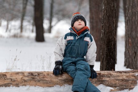 Foto de El chico se sienta pensativamente en el tronco en el bosque de invierno. El niño se sienta con las piernas cruzadas y mira al cielo - Imagen libre de derechos