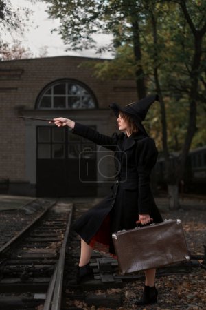 Photo for Girl dressed in black cape, pointed hat and holds retro suitcase in hands. Young witch with magic wand conjures at old train station. Vertical frame - Royalty Free Image