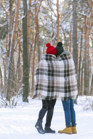 Photo for Young man and woman standing embracing and wrapped in a plaid in a snowy winter forest. Back view. Copy space. - Royalty Free Image