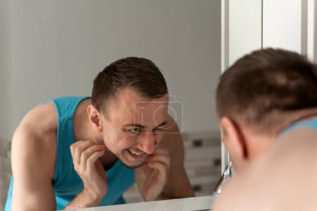 Photo for Middle-aged man with no shaving face stands bent over sink, unpleasure looks at reflection in mirror and scratch his bristle. Morning hygiene. - Royalty Free Image