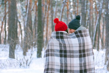 Photo for Young man and woman sitting embracing and wrapped in a plaid in a snowy winter forest. Back view. Copy space. - Royalty Free Image