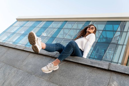 Photo for Attractive stylish woman resting on city street.Young brunette girl sits on parapet with her legs hanging down.Urban. - Royalty Free Image