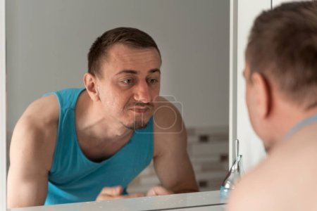 Photo for Middle-aged man with no shaving face stands bent over sink and unpleasure looks at reflection in mirror. Morning hygiene. - Royalty Free Image
