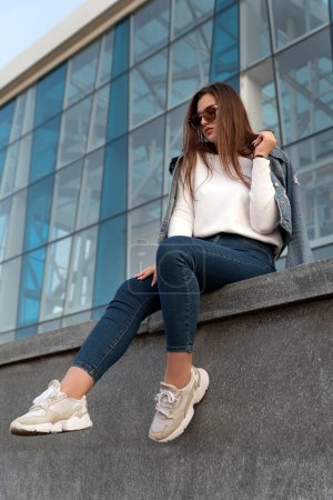 Photo for Portrait of stylish young woman in sunglasses on city street. Attractive woman resting next to modern building. Vertical. - Royalty Free Image