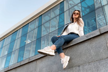 Photo for Attractive brunette girl sits on parapet with her legs hanging down. Urban. Stylish woman resting on city street. - Royalty Free Image