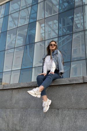 Photo for Young woman in sunglesses in youth clothing on the city street. Attractive stylish woman resting on city street. Vertical. - Royalty Free Image