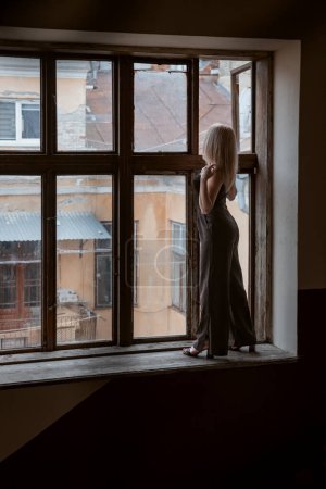 Photo for Young woman in black pantsuit stands on the windowsill of large window and looks at the building. Vertical frame - Royalty Free Image