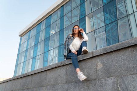 Photo for Sexy young woman in fashionable youth clothing and sunglasses resting on the city street. Girl student on parapet. - Royalty Free Image