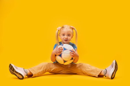 Little blonde girl with two tails sits on floor and holds soccer ball on yellow background. Womens childrens football